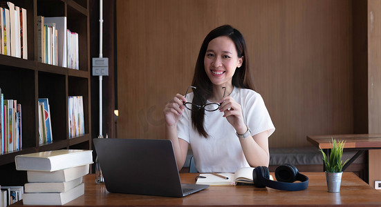出国留学亚洲学生戴着眼镜看着相机桌子上有一台笔记本电脑可以使用朋友的互联网连接进行国际视频通话图书馆里灿烂的笑容。