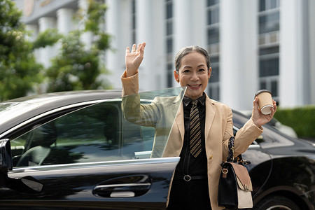友好的亚洲高级女士打招呼，挥舞着手，带着幸福的微笑问候你，下了她的豪华车。