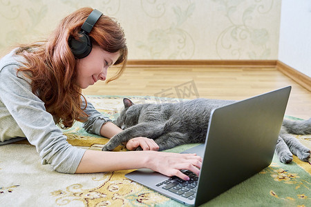 猫地板摄影照片_戴着耳机、笔记本电脑和睡觉的灰猫的青春期前女孩