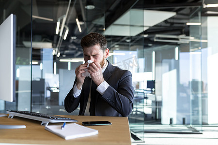一个流鼻涕和发烧的男人在办公室工作，一个生病的商人在电脑前流鼻涕