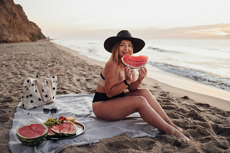 清晨坐在海边沙滩上的格子上，年轻漂亮的女人拿着西瓜片