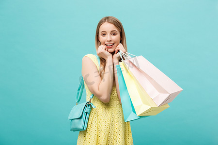 蓝色购物摄影照片_生活方式概念：穿着黄色夏季服装、拿着购物袋、在蓝色背景下看着相机的震惊的年轻漂亮女人的肖像。