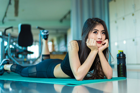 运动馆背景摄影照片_运动女子躺在有运动器材背景的健身馆的瑜伽垫上放松。