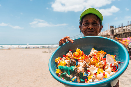 海滩上年长的拉丁裔街头小贩展示了一碗用彩绘贝壳制成的手工艺品。