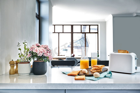 面包鸡蛋橙汁摄影照片_浅色厨房桌上的烤面包机，配有新鲜面包、鸡蛋和一杯橙汁