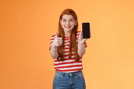 快乐满意的红发迷人女孩欢呼展示拇指，推荐智能手机应用程序，展示移动屏幕，介绍设备，批准好礼服展示网上商店手机，橙色背景