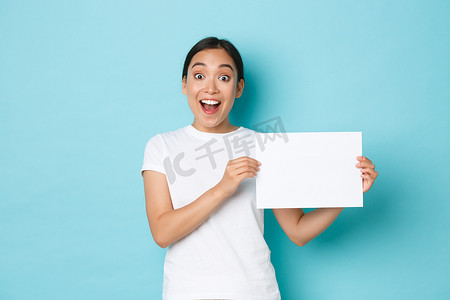 穿着白色 T 恤的亚洲女孩惊奇而兴奋，发布公告，在展示标志时惊奇地喘着粗气，一张空白纸上贴着你的广告，站在浅蓝色背景中