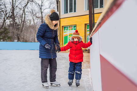 教室外摄影照片_年轻的母亲教她的小儿子在室外溜冰场溜冰。