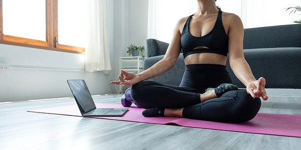 手部妇女在瑜伽垫上做莲花瑜伽练习的特写，早上在家里的平板电脑上进行在线训练，放松和冥想的概念