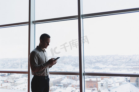 一位年轻的高管站在办公室内部，拿着电话，透过一扇俯瞰城市的大窗户往外看。