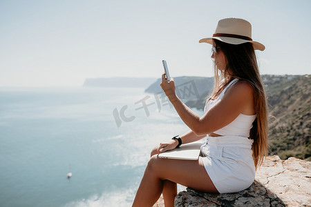 坐在海边的女摄影照片_数字游牧者，戴帽子的女人，一位带着笔记本电脑的女商人在日落时坐在海边的岩石上，从远处进行在线商业交易。