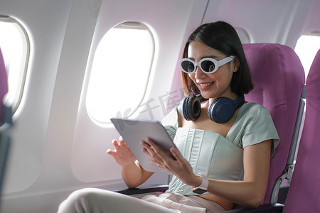 旅游小程序摄影照片_旅游与现代技术和航空概念，女性坐在飞机上与现代数字小工具和搜索最喜欢的音乐播放列表在应用程序中聆听