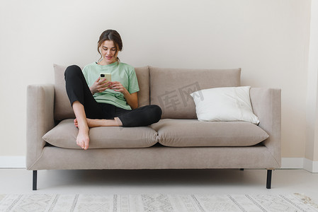 快乐乐观的年轻女子坐在家里的室内，在家里的沙发上使用手机购物或社交媒体的形象。
