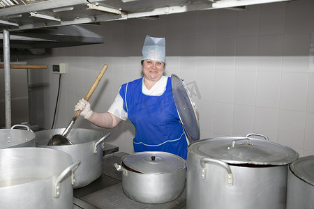 学雷锋做好事摄影照片_一位女厨师在工业厨房里做饭，金属锅中放着一个大勺子。