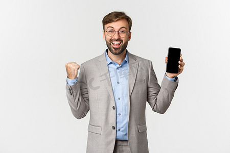 穿着灰色西装和眼镜的快乐商人，在移动应用中实现日常目标，展示智能手机屏幕并欢欣鼓舞，赢得一些东西，站在白色背景上