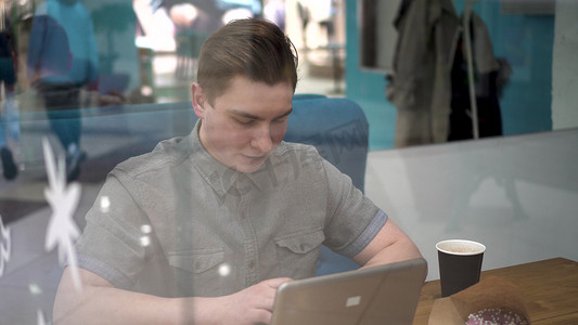 咖啡馆摄影照片_年轻人坐在咖啡馆的玻璃后面，带着笔记本电脑。