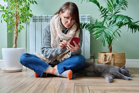 在家使用智能手机的女性，在暖气片附近用猫取暖