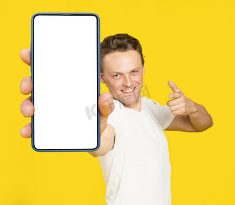 巨大手机摄影照片_拿着巨大的智能手机与白色屏幕英俊的金发男子指着白色空屏幕，穿着白色 T 恤和牛仔裤隔离在黄色背景。