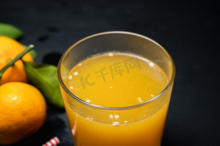 黑暗的表面上有三个橘子，叶子旁边有一杯果汁。