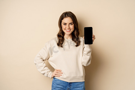 界面购物摄影照片_显示智能手机屏幕、移动应用程序界面、站在米色背景上的时尚微笑女性