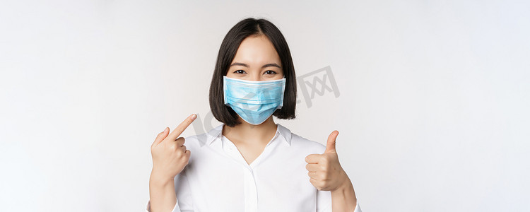 戴着医用面具的亚洲女孩的画像竖起大拇指，指着她的 covid 保护，站在白色背景上