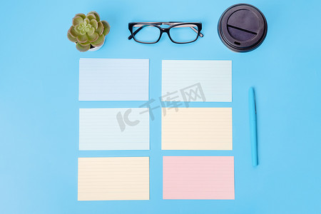 蓝色背景上的空白、信纸、纸、咖啡、眼镜、植物和笔。