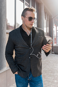 一位身穿太阳镜、夹克和衬衫、牛仔裤和耳机的年轻商人的肖像，在户外街上用手机听音乐