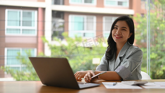 坐着的会计摄影照片_漂亮的亚洲女经理坐在明亮的现代办公室的笔记本电脑前，对着镜头微笑