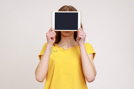 身着黄色休闲风格 T 恤的未知女性肖像，脸上覆盖着数字平板电脑，屏幕空空，广告复制空间。