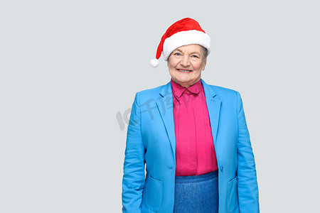 穿着蓝色西装、戴着圣诞圣诞老人红帽的老年妇女站着看着相机