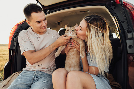 年轻男女带着宠物坐在汽车后备箱里，幸福的夫妇带着红猫享受公路旅行