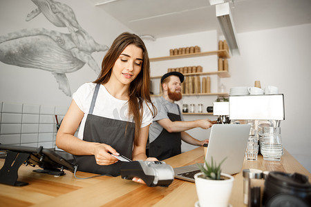 咖啡经营理念-美丽的女咖啡师在现代咖啡店的吧台工作时用信用卡为顾客提供付款服务并微笑。