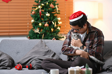戴着圣诞老人帽子的年轻亚洲男子和他可爱的猫坐在圣诞树附近的沙发上。