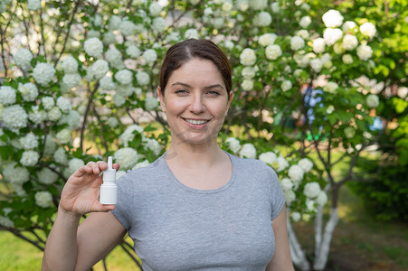 幸福的女人在一棵开花的树的背景下展示鼻喷雾剂。