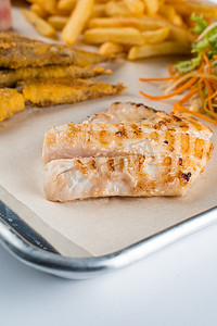 白鱼摄影照片_在沙拉、炸薯条背景的烤鱼片白鱼，在金属板上。