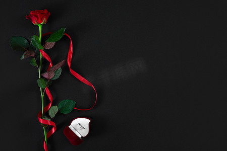 一朵深红色的玫瑰，上面有红色丝带和黑色背景上有戒指的盒子