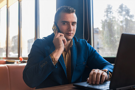 一位商人坐在窗边咖啡馆的桌子旁，穿着漂亮的欧洲西装，用笔记本电脑工作，用手机或手机通话，让商人感到惊讶