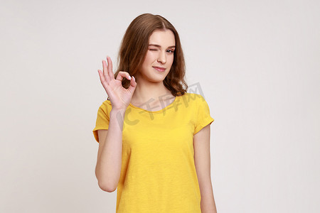 满意的年轻女子的肖像，她留着棕色波浪发，穿着休闲黄色 T 恤，站着，看着镜头，摆出“OK”手势并眨眼。