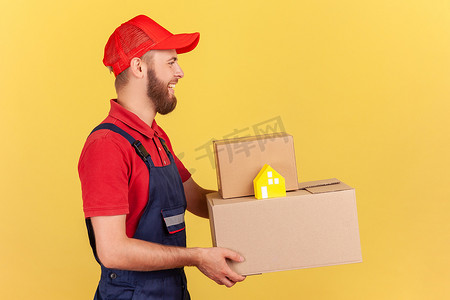 红帽摄影照片_身穿制服、头戴红帽的送货员拿着纸箱和纸屋，送货上门。