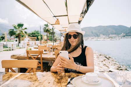 戴着墨镜的年轻女女士在街头海滨咖啡馆餐厅咖啡店里看着她的移动智能手机小工具，背景是风景山。