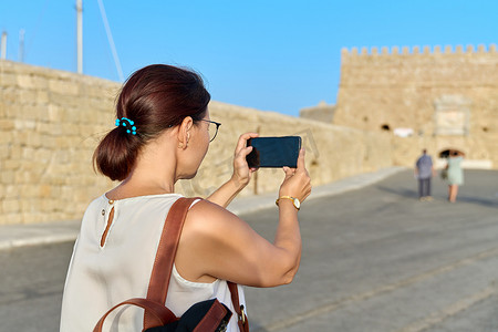 海滨城市图片摄影照片_女游客用智能手机拍照，这是海滨的一座古老历史堡垒