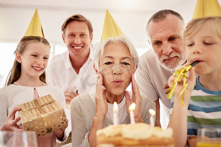高级女士在家里与家人一起庆祝生日，戴着派对帽，吹着口哨。