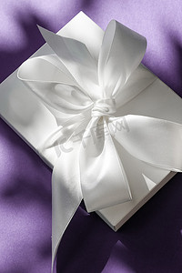 绸带丝带飘带摄影照片_紫色背景、奢华婚礼或生日礼物上带丝带和蝴蝶结的豪华假日白色礼盒