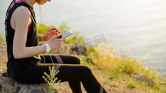 路上听摄影照片_年轻女子跑步者使用智能手机，在日落时分在山路上听音乐。