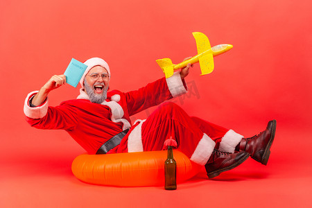 兴奋的留着灰色胡子的老人穿着圣诞老人服装坐在橡皮圈上，拿着纸飞机和护照，享受冬季旅行。