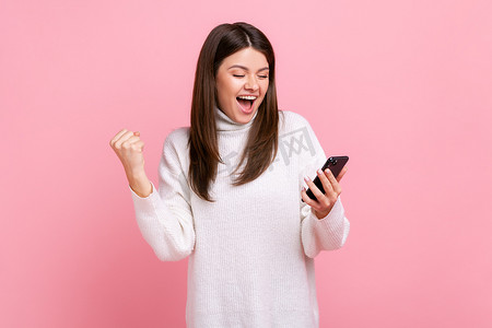 快乐的积极女孩手里拿着智能手机,握紧拳头,庆祝胜利,突然的成功.
