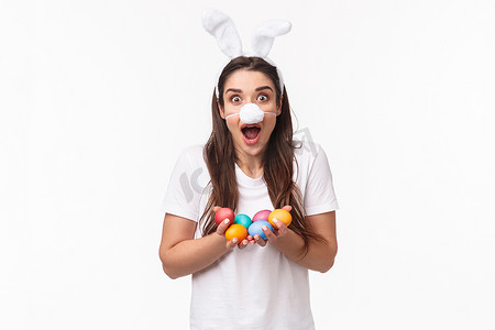 拿着彩蛋摄影照片_穿着兔子服装、耳朵和蓬松鼻罩、拿着彩蛋、庆祝节日复活节派对的兴奋快乐的年轻风趣女人的肖像，令人惊讶的白色背景
