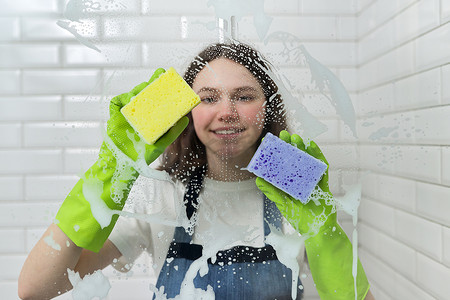 浴室玻璃摄影照片_浴室清洁，少女用泡沫和海绵清洗淋浴玻璃