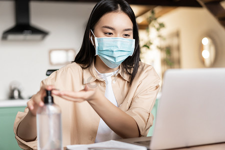 上洗手摄影照片_戴着医用面罩的亚洲年轻女性坐在联合办公空间，在笔记本电脑上参加在线讲座或网络研讨会，并使用洗手液洗手