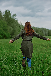 春天黄昏摄影照片_春天阴天，一位穿着长雨衣的女人跑过高高绿草的田野